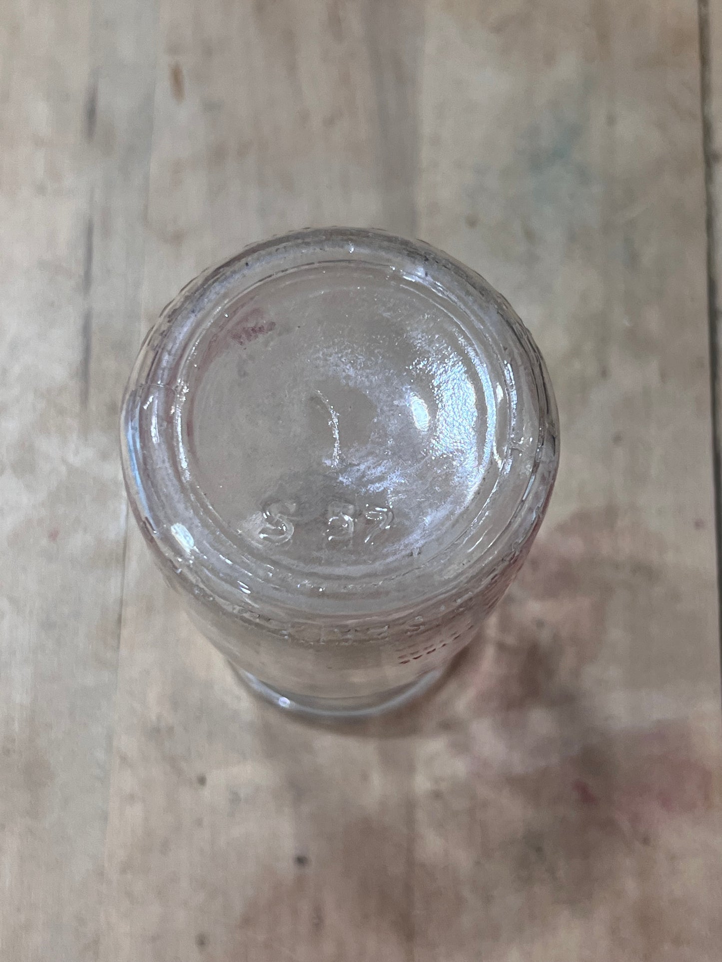 A clear glass jar or bottle viewed from above, showing the bottom of the container with embossed markings, placed on a light wood surface.