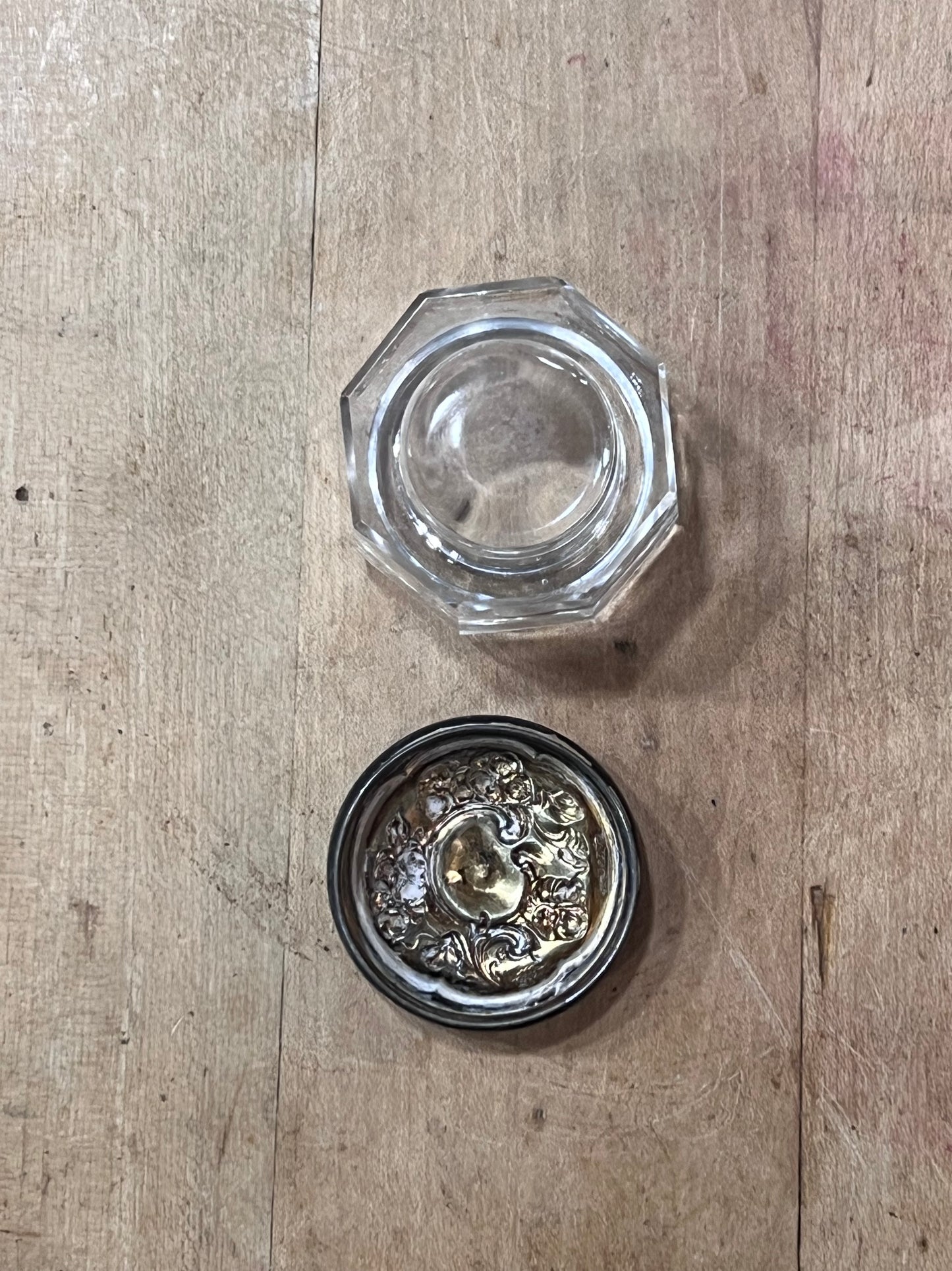 A clear glass octagonal container with its silver-toned ornate lid placed below it on a light wood surface.