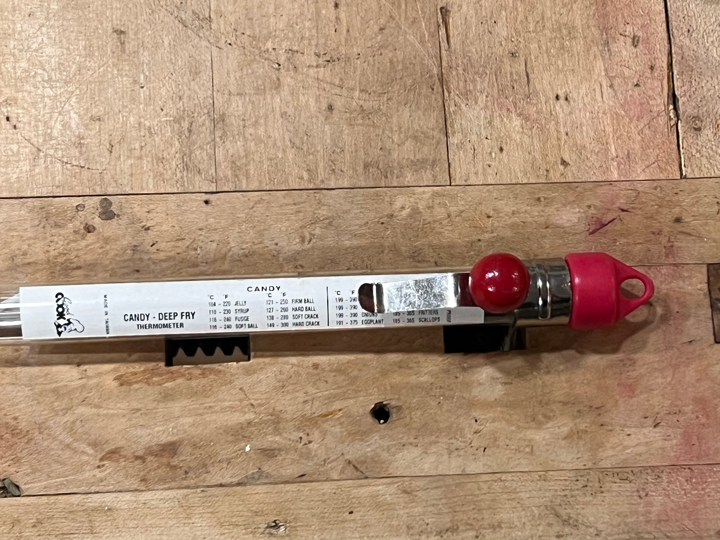 A candy thermometer tool with a white printed scale label, a red spherical bulb, and a red cap, resting on a wooden board.