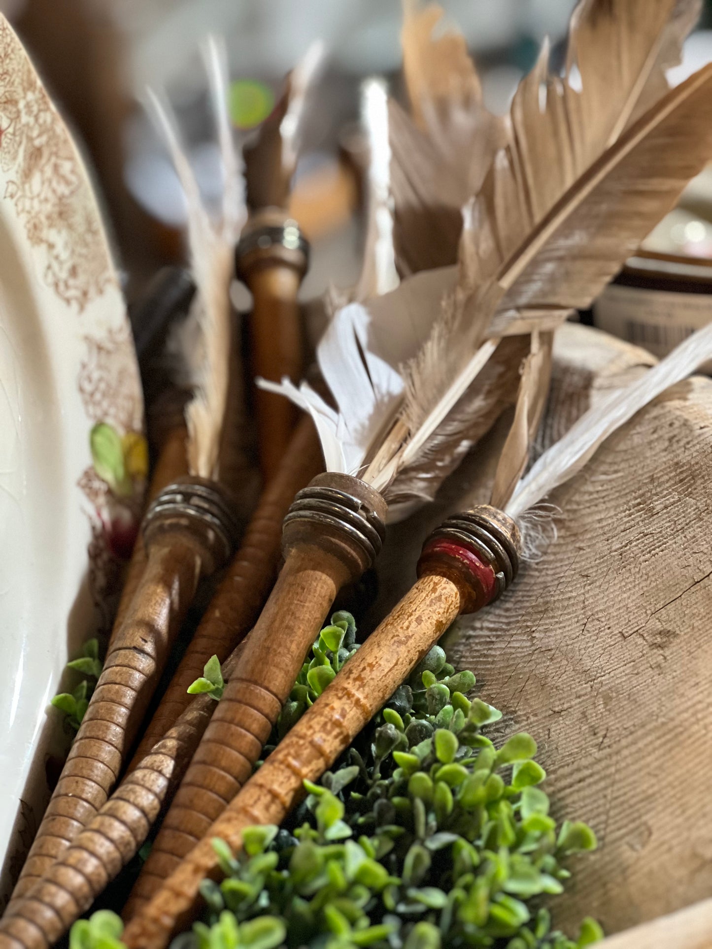 A close-up of several decorative feather quills with ribbed wooden handles and metallic accents, arranged with small green foliage on a rustic wooden surface.