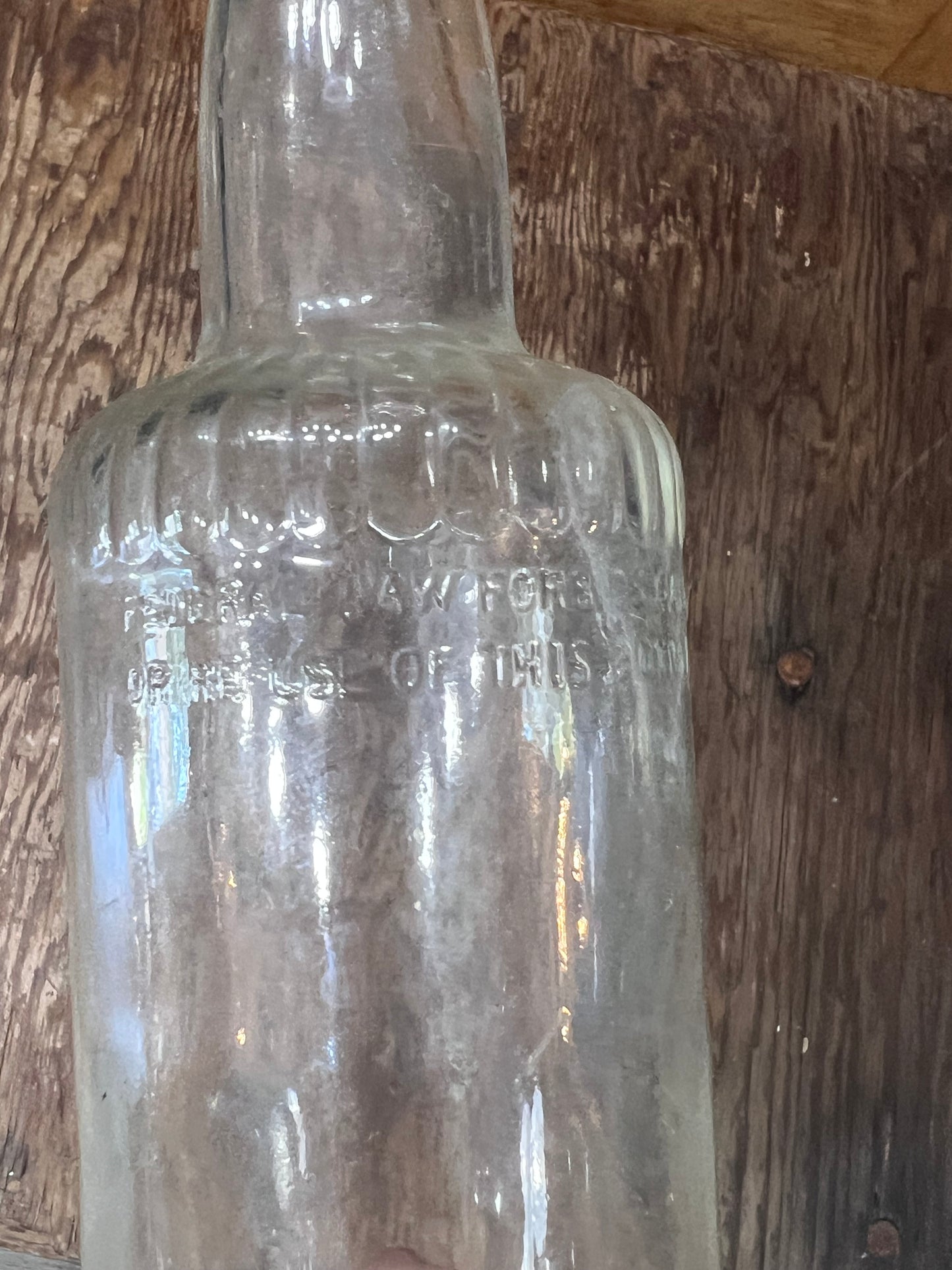 A clear vintage glass bottle with embossed text, shown against a rustic wooden plank background.