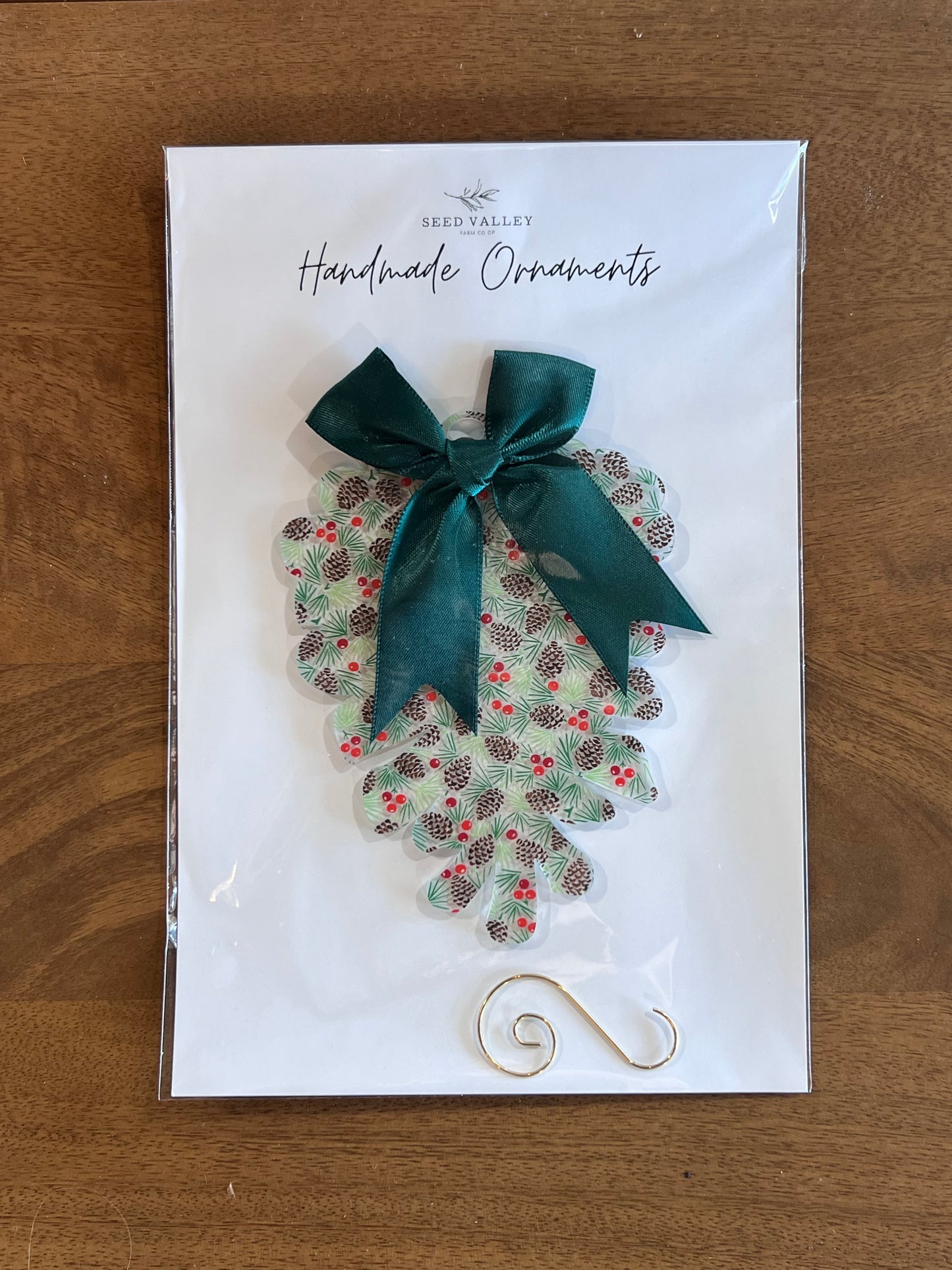A Seed Valley handmade ornament displayed on a white backing card, featuring a scalloped green and brown patterned ornament with a dark green satin bow, placed on a wooden surface.