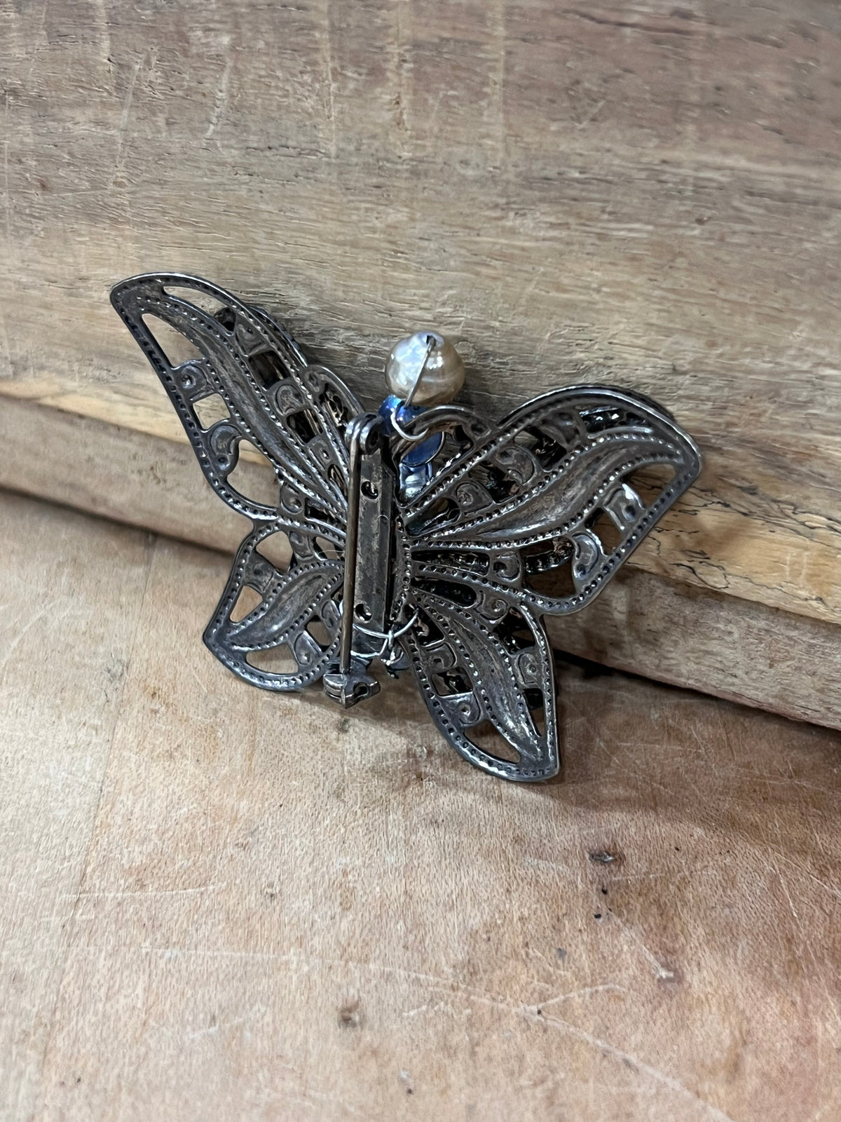 A butterfly-shaped filigree brooch in dark oxidized silver metal, featuring a small clear faceted crystal at the center, resting on a rustic wooden surface.