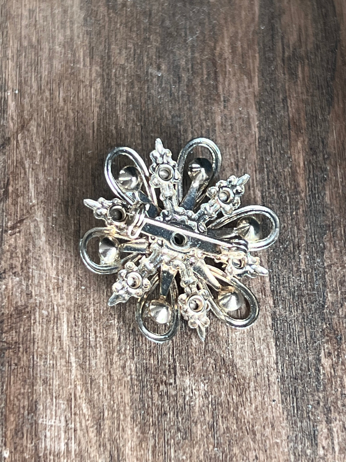 An ornate silver-toned metal brooch with a vintage floral or scroll design, displayed on a dark brown wooden surface.