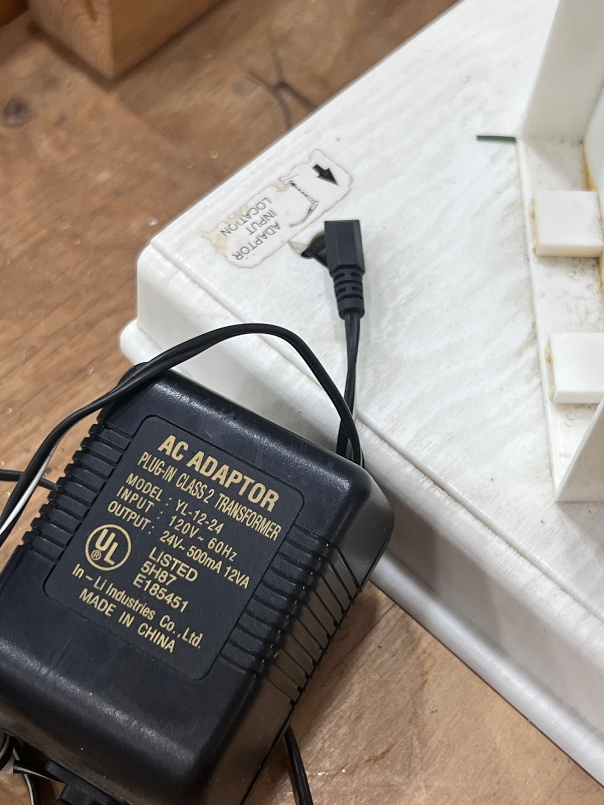 A black AC adapter power supply with gold text, shown next to a white plastic case on a wooden surface.