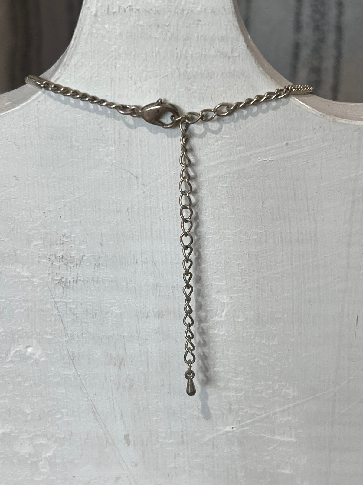 A silver-toned chain necklace displayed on a white textured mannequin bust, featuring a lobster clasp closure and a long dangling heart-link extender chain.