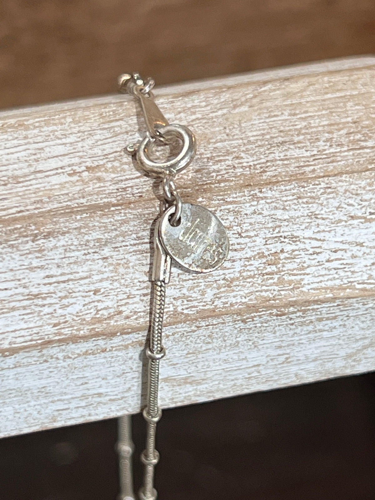 Close-up of a silver-toned chain bracelet with a lobster clasp, ring, oval charm, and a small round tag resting on a distressed white wooden surface.