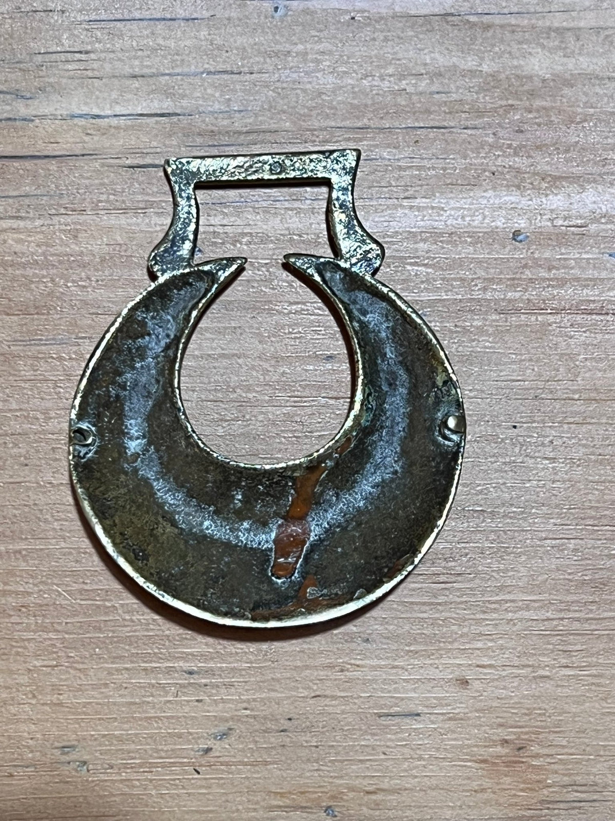 A vintage metal horseshoe-shaped pull tab or decorative hardware piece with a rectangular top loop, shown on a light brown wooden surface.