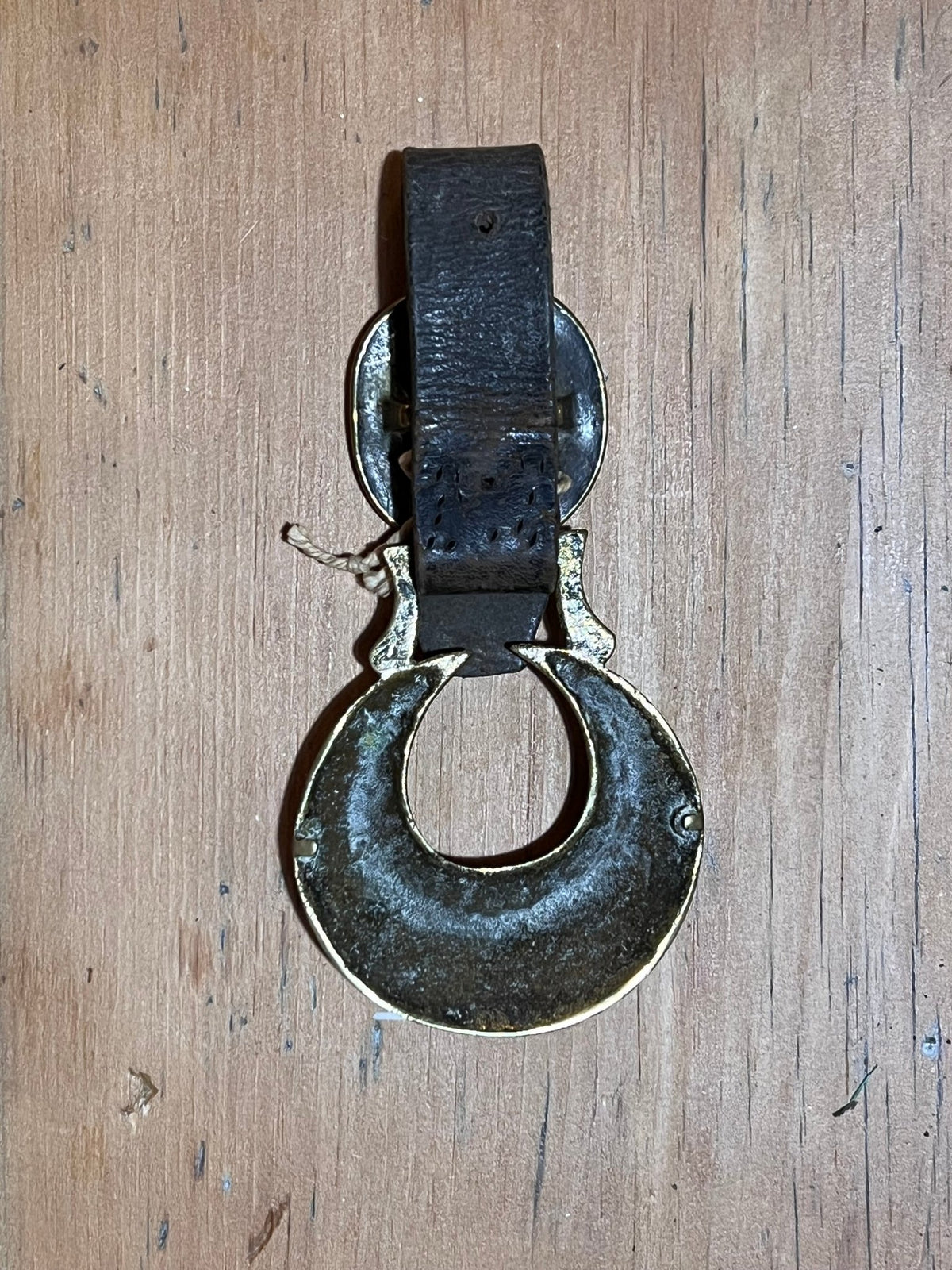 A vintage dark metal ring-shaped door pull with a black leather strap, displayed on a light brown wooden surface.