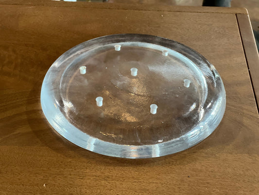 A clear, round acrylic or glass trivet with six small raised feet, shown on a wooden tabletop.