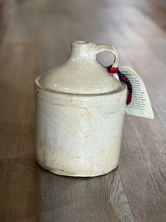 A handmade ceramic stoneware jug with a lid and handle, featuring a speckled cream glaze and a small red and black ribbon tag attached, set on a wooden floor.