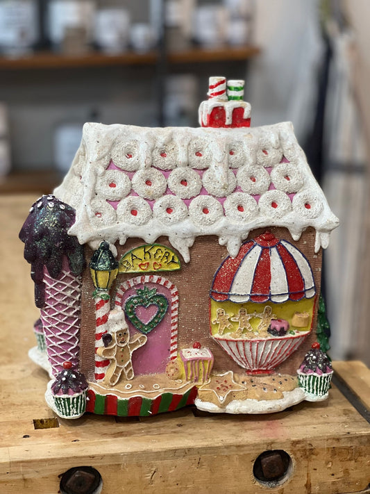 A decorative gingerbread-style house ornament with a candy shop theme, featuring a white icing-draped roof, pink door, striped awning, and cupcake details, displayed on a wooden surface.