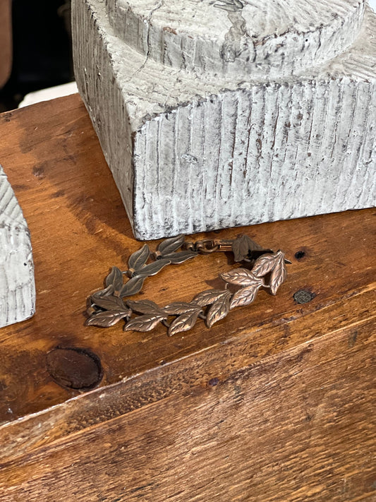 A bronze-toned leaf wreath bracelet resting on a rustic wooden table next to a textured gray stone block.
