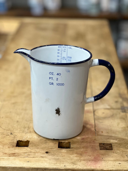 A vintage white enamel measuring pitcher with a dark blue rim, handle, and printed measurement markings, resting on a rustic wooden workbench.