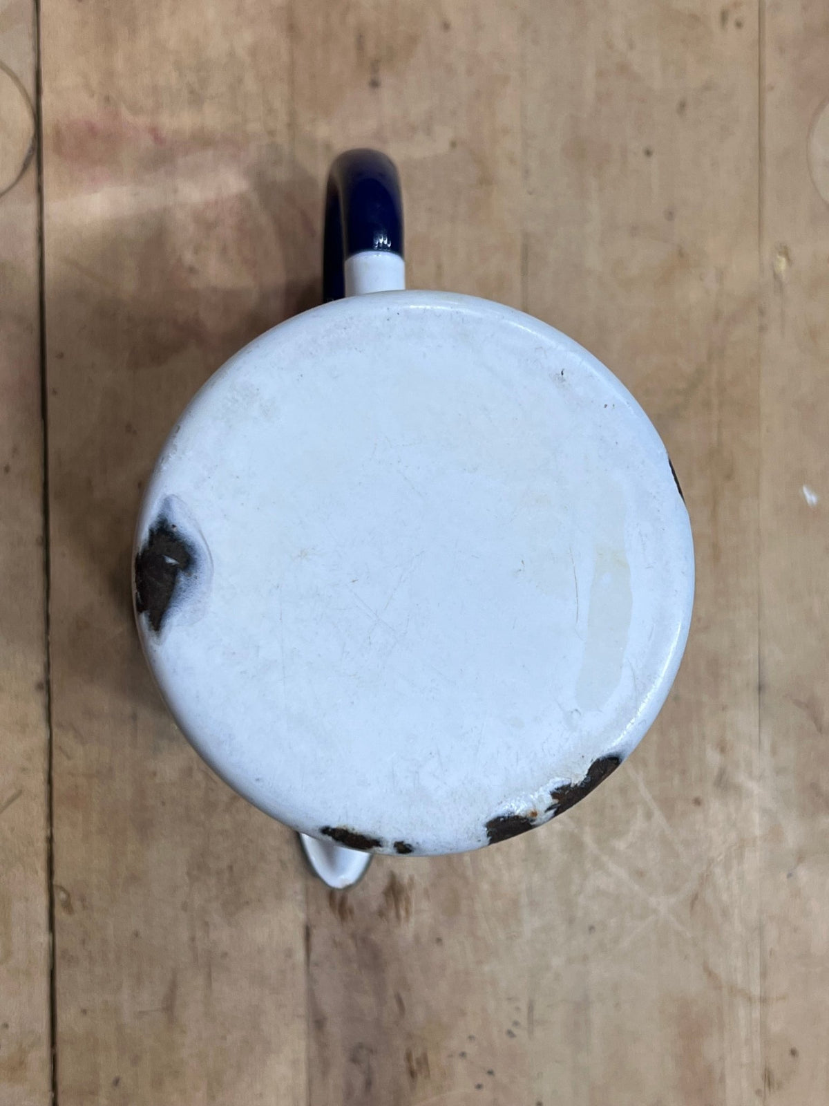 A top-down view of a vintage white enamel mug with a dark blue handle, resting on a light brown wooden surface.
