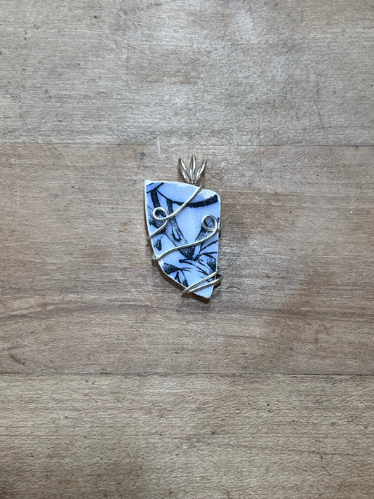 A handmade pendant featuring a light blue and white patterned stone wrapped in silver-colored wire, displayed on a rustic wooden surface.