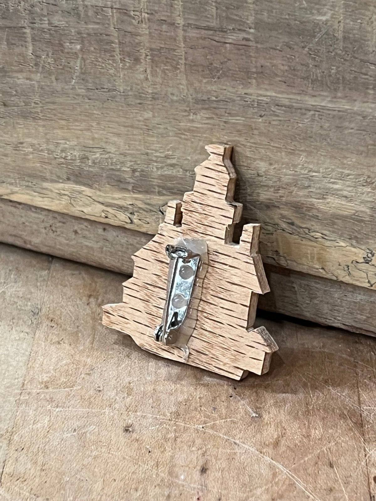 A small wooden Christmas tree-shaped brooch with a silver metal pin back, resting on a rustic wooden surface.