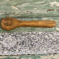A rectangular gray and white speckled granite stone slab with a circular hole, shown next to a small hand-carved wooden spoon engraved with 'Taste tester' on a distressed green wooden background.