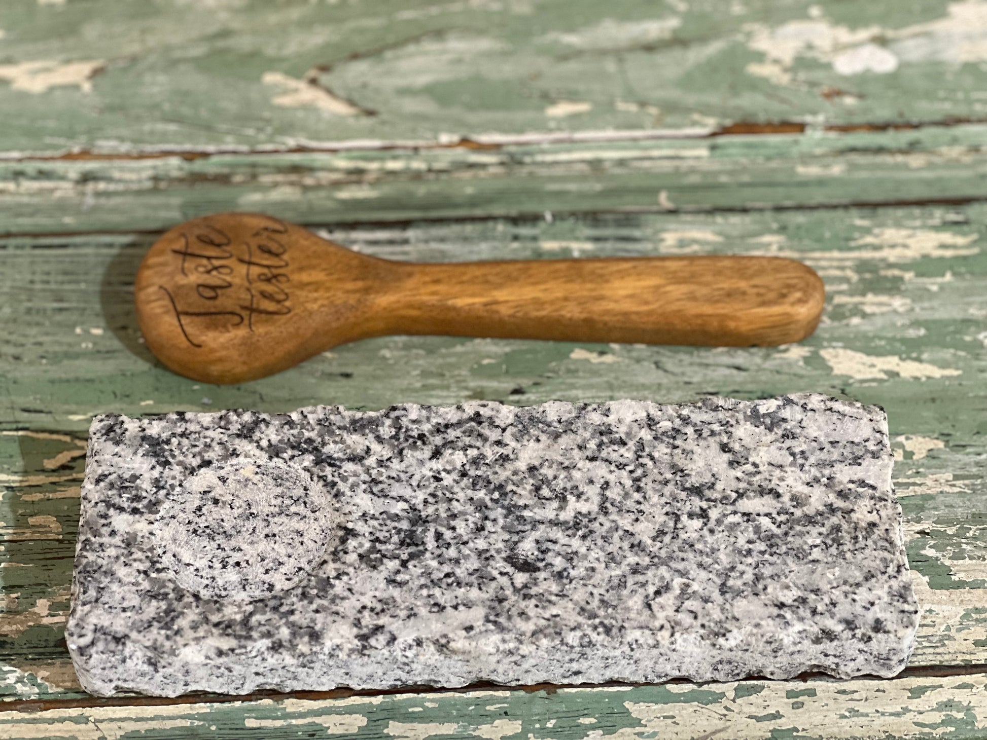 A rectangular gray and white speckled granite stone slab with a circular hole, shown next to a small hand-carved wooden spoon engraved with 'Taste tester' on a distressed green wooden background.