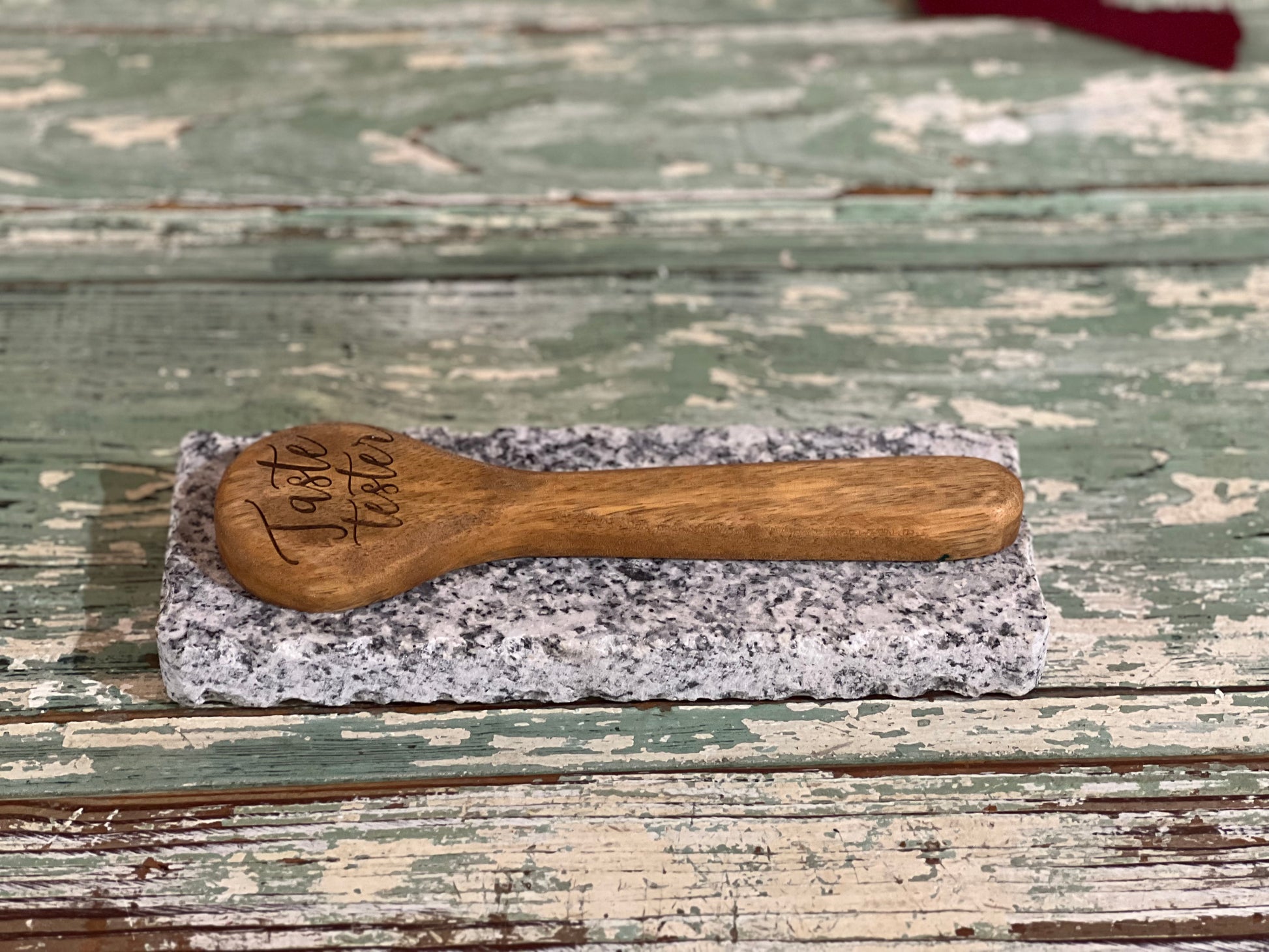 A small wooden spoon engraved with 'Taste Better' rests on a rectangular gray stone slab over a distressed green wooden surface.