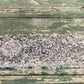 A rectangular gray granite stone block with a circular cutout, displayed on a rustic, weathered green wooden surface.