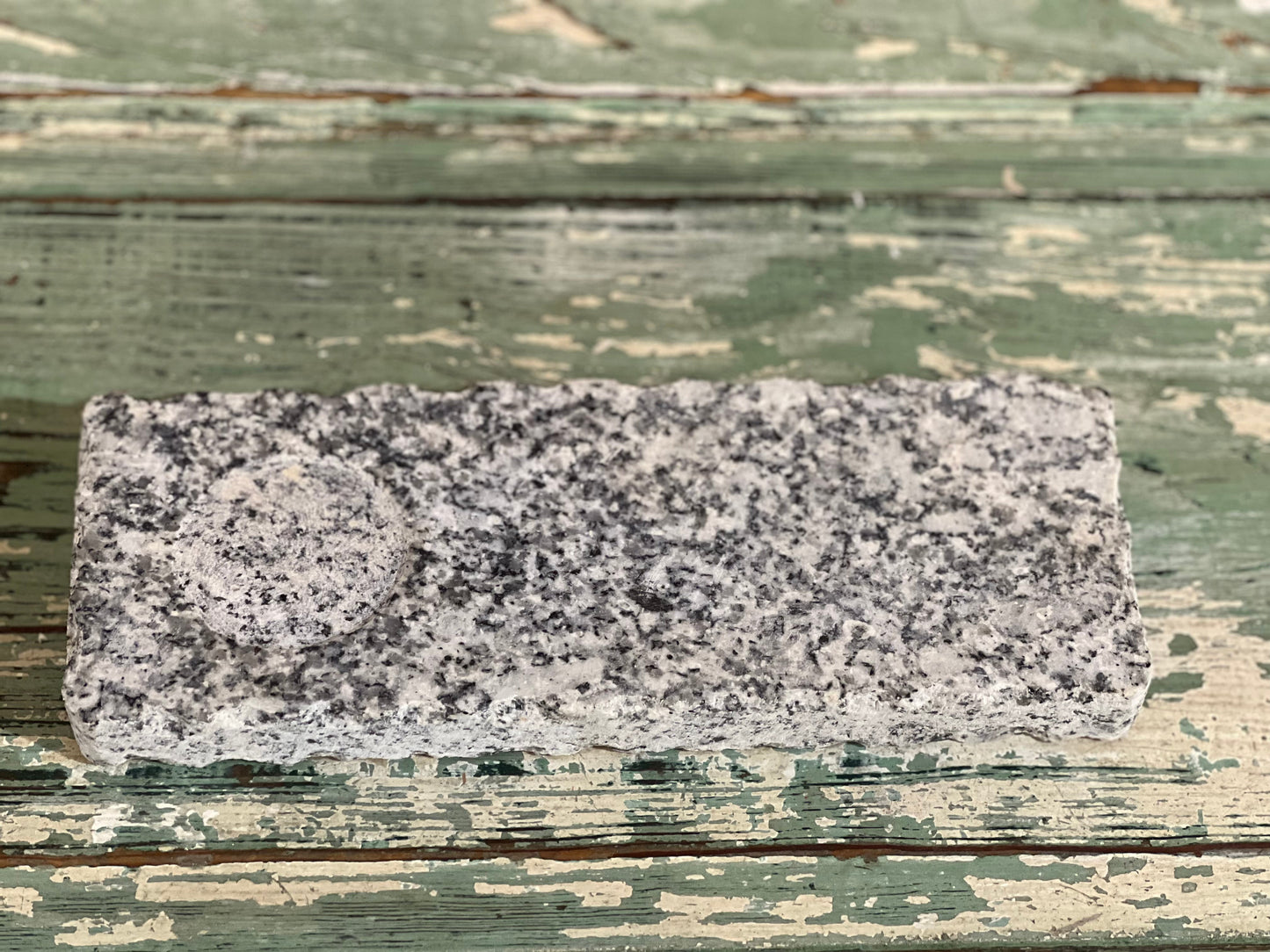 A rectangular gray granite stone block with a circular cutout, displayed on a rustic, weathered green wooden surface.