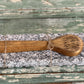 A small handcrafted wooden spoon with 'Faith Farrow' engraved on the bowl, wrapped in twine and resting on a rectangular gray-and-white speckled stone slab over a distressed green wooden surface.