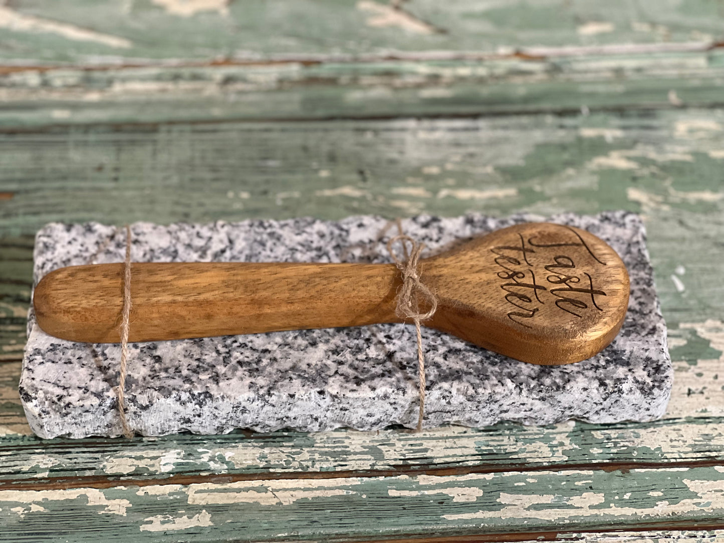 A small handcrafted wooden spoon with 'Faith Farrow' engraved on the bowl, wrapped in twine and resting on a rectangular gray-and-white speckled stone slab over a distressed green wooden surface.
