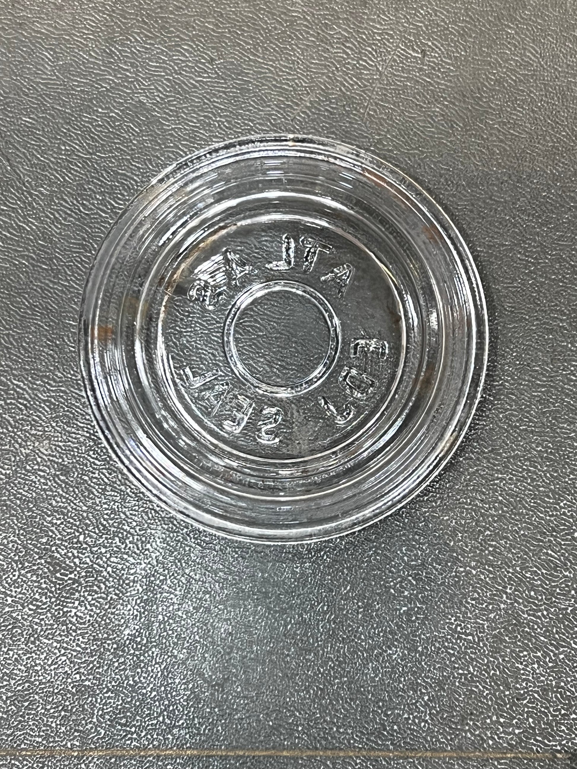 A clear glass round shallow dish or saucer with embossed text reading 'VINTA 500' on a textured gray surface.