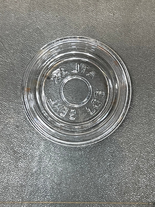 A clear glass round shallow dish or saucer with embossed text reading 'VINTA 500' on a textured gray surface.