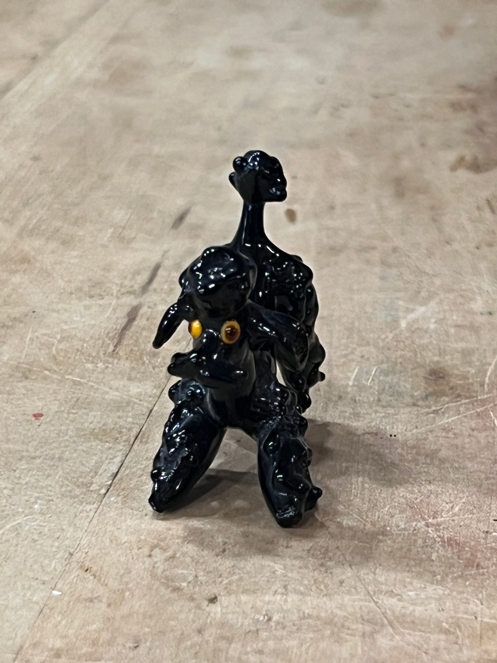 A small glossy black figurine of a dog with yellow eyes, standing on a scratched light brown wooden surface.