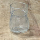 A clear glass cup with a flared rim and ribbed body, lying on a light-colored wooden surface.