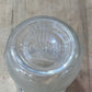 A clear glass jar viewed from above, showing the embossed brand name 'Oster' on the glass base, resting on a light-colored wooden surface.