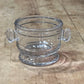 A clear glass bowl with decorative scalloped handles, shown on a rustic wooden surface.