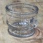 A clear glass jar with a scalloped, decorative rim and a small matching glass knob on top, resting on a textured, light brown wooden surface.