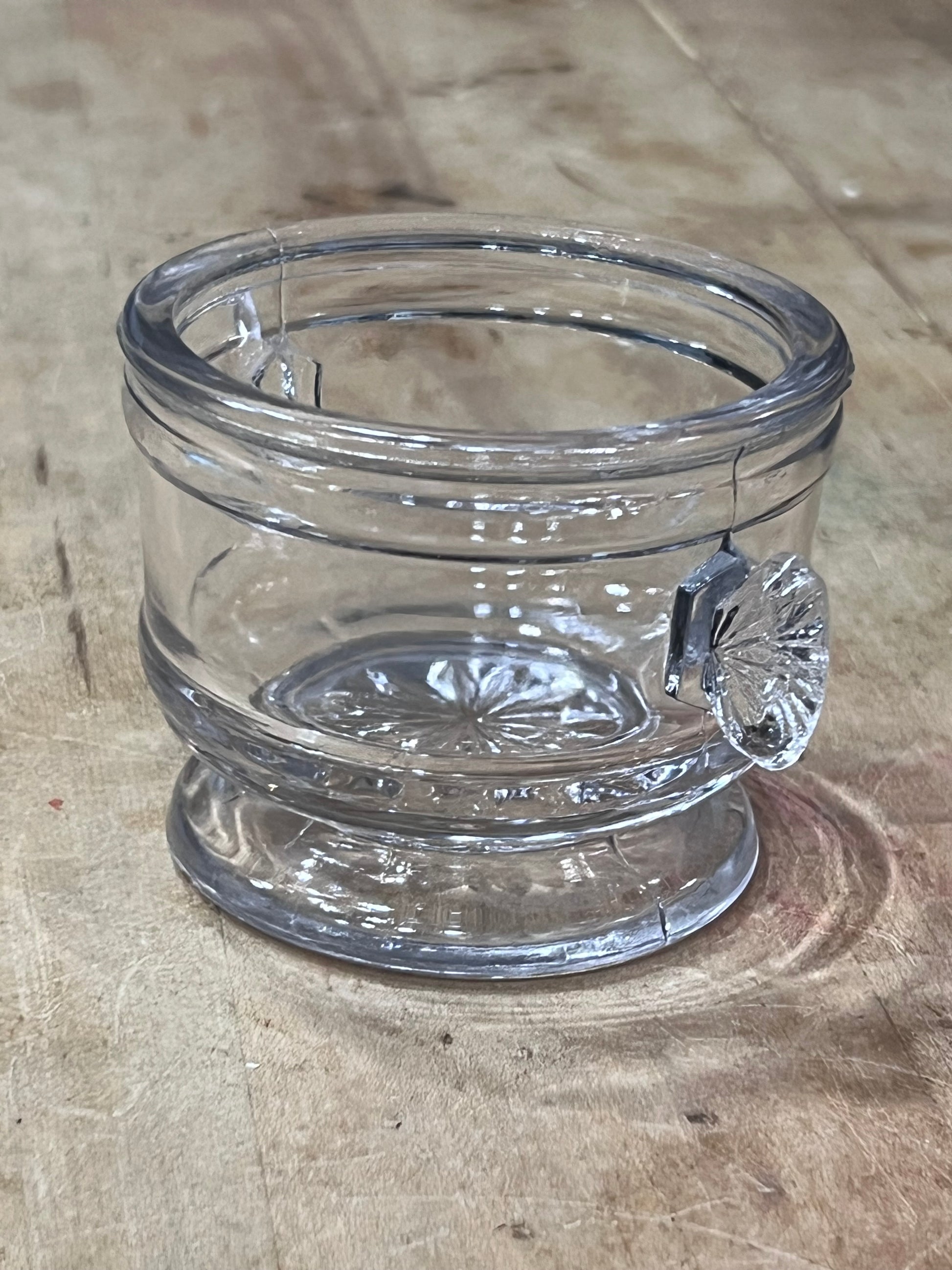 A clear glass jar with a scalloped, decorative rim and a small matching glass knob on top, resting on a textured, light brown wooden surface.