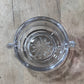 A clear glass ashtray with a decorative starburst pattern in the center and small hanging tabs, shown from above on a wooden surface.