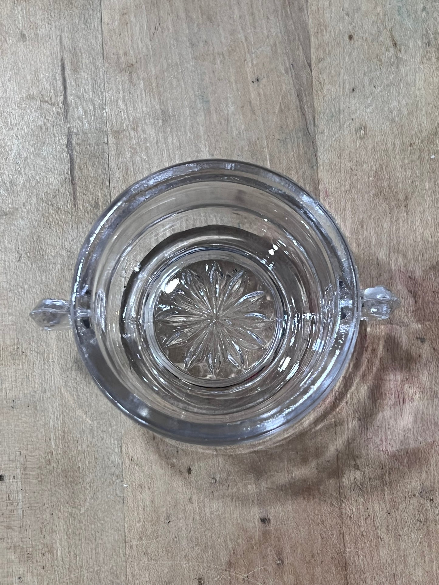 A clear glass ashtray with a decorative starburst pattern in the center and small hanging tabs, shown from above on a wooden surface.
