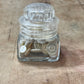 A small clear glass jar with a ribbed screw-top lid, shown on a worn wooden surface. Inside the jar are several off-white buttons.