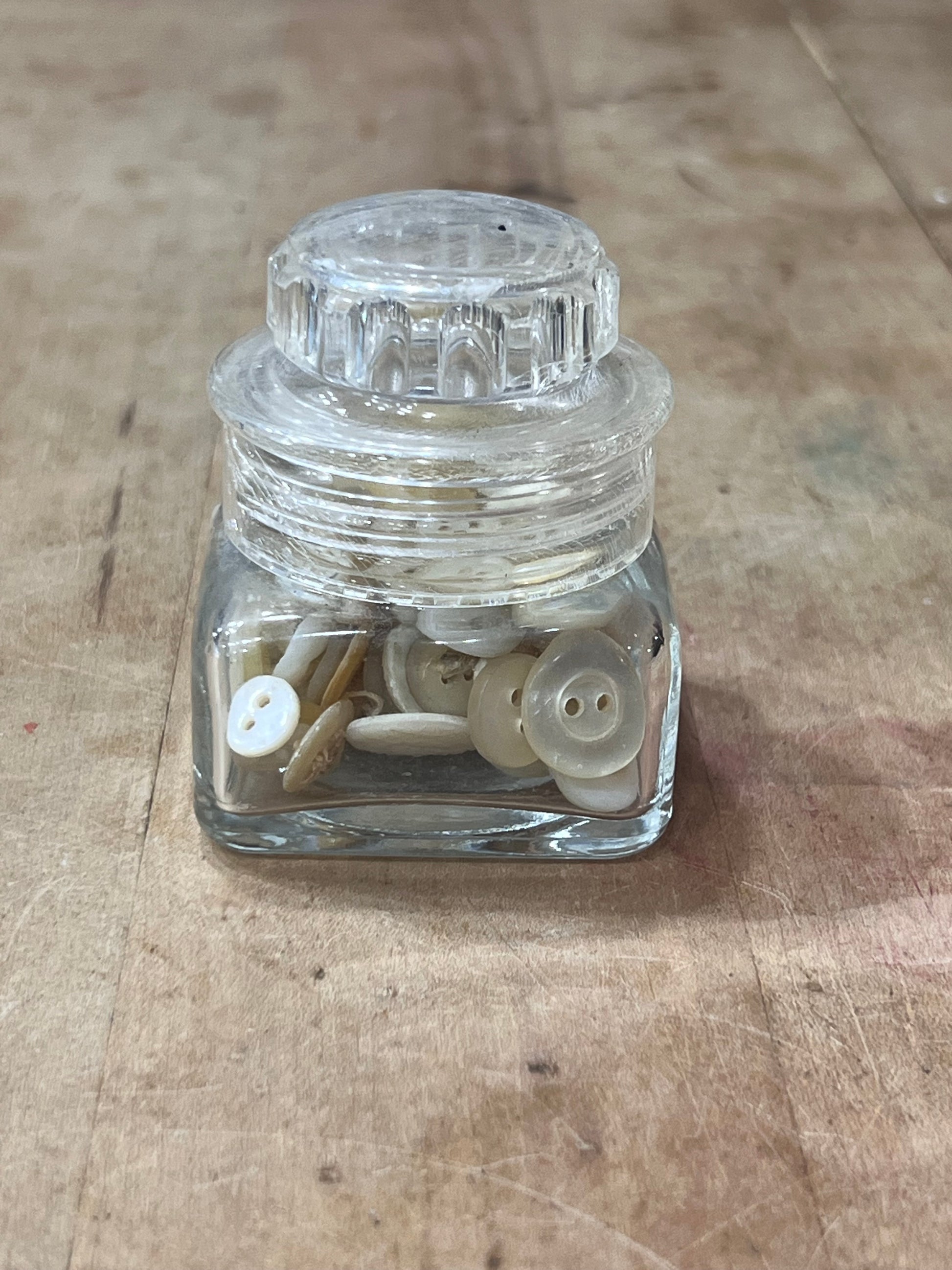 A small clear glass jar with a ribbed screw-top lid, shown on a worn wooden surface. Inside the jar are several off-white buttons.