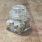 A small clear glass hexagonal jar with a ribbed screw-top lid, tipped on its side and filled with white buttons, placed on a worn wooden surface.