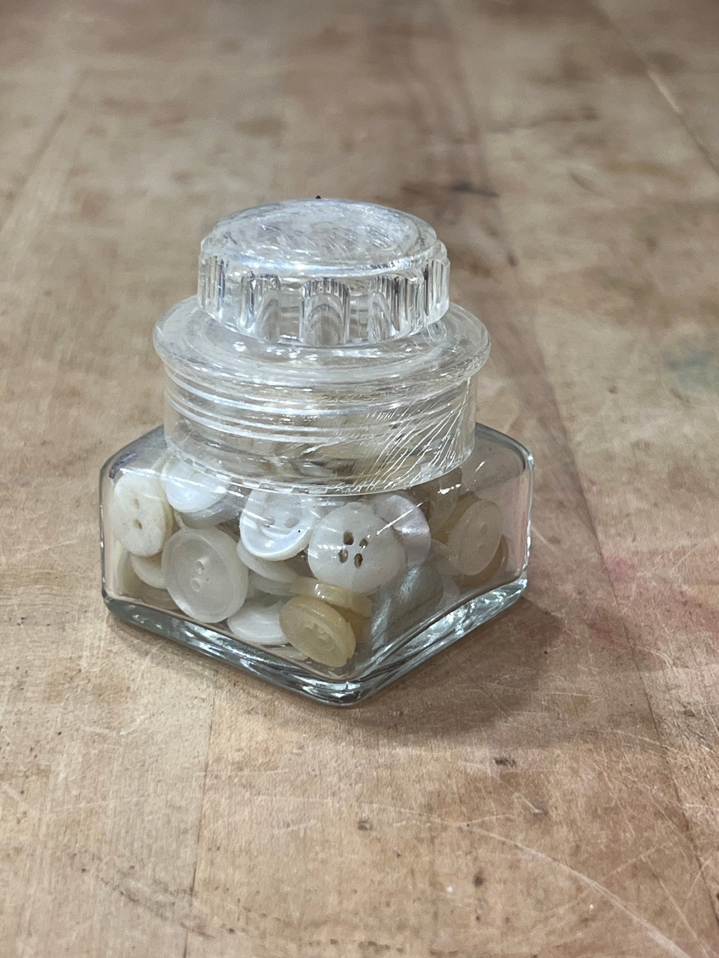 A small clear glass hexagonal jar with a ribbed screw-top lid, tipped on its side and filled with white buttons, placed on a worn wooden surface.