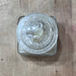 A clear glass jar with a square base, shown from a top-down view on a light brown wooden surface. Inside the open jar are several small white buttons.