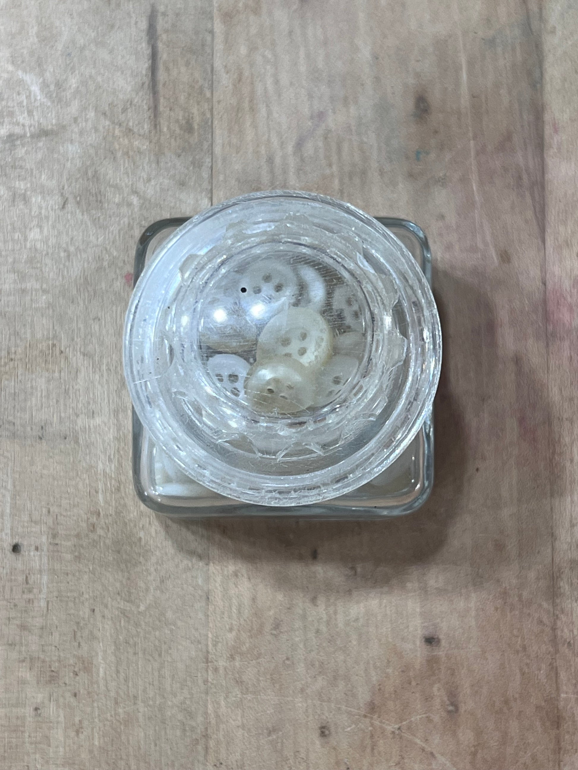 A clear glass jar with a square base, shown from a top-down view on a light brown wooden surface. Inside the open jar are several small white buttons.