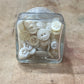 A small clear glass container filled with assorted off-white and cream buttons, photographed from above on a light wood surface.