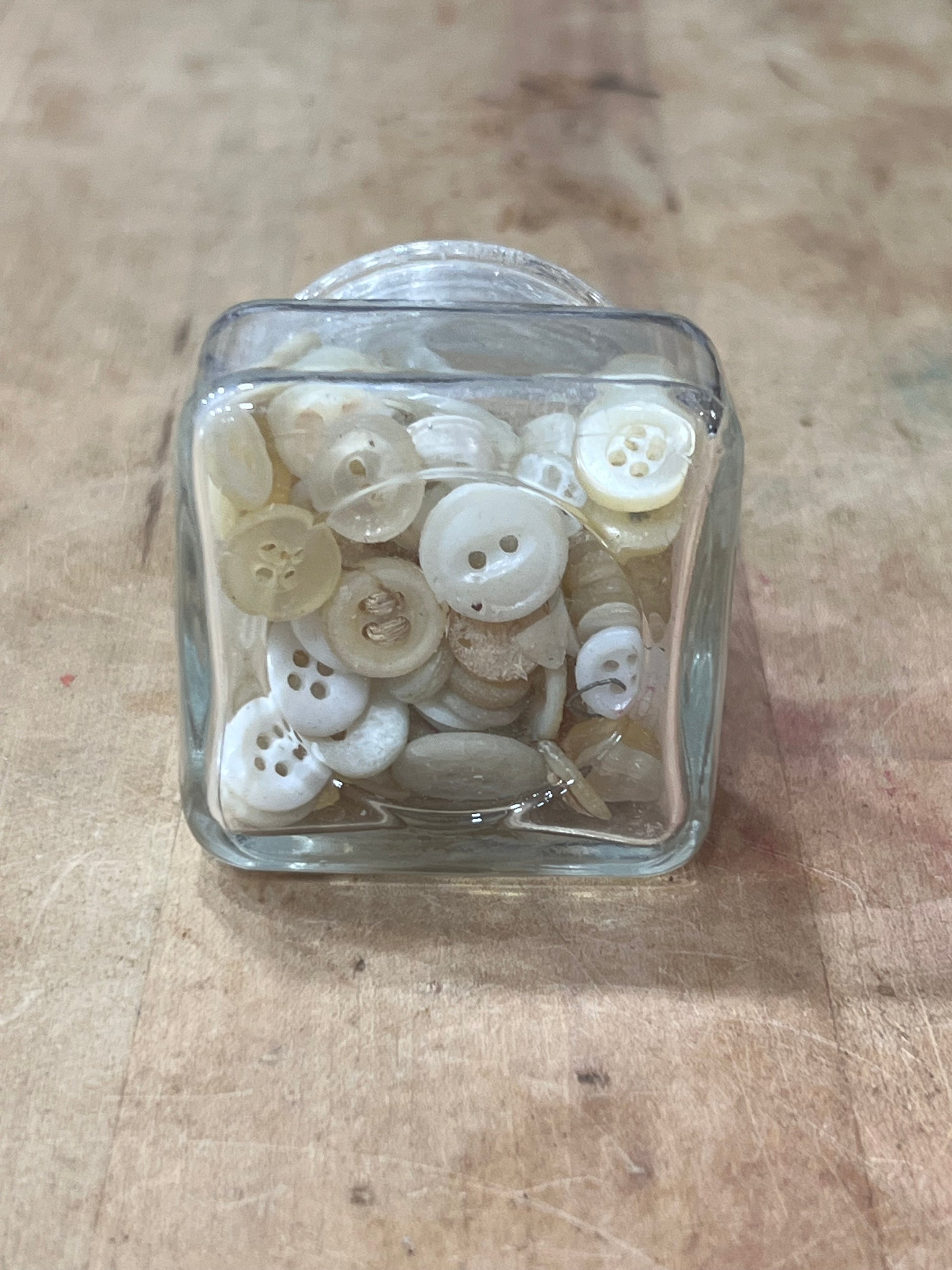 A small clear glass container filled with assorted off-white and cream buttons, photographed from above on a light wood surface.