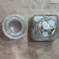 A small clear glass jar filled with white buttons next to its matching clear glass lid, placed on a rustic wooden surface.