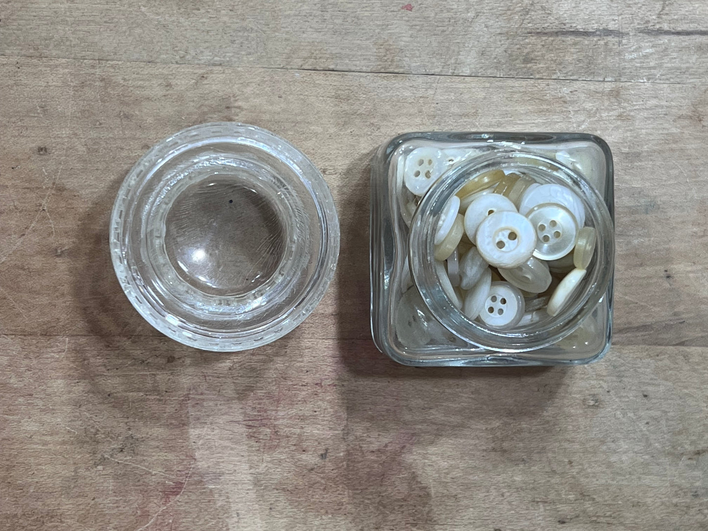 A small clear glass jar filled with white buttons next to its matching clear glass lid, placed on a rustic wooden surface.