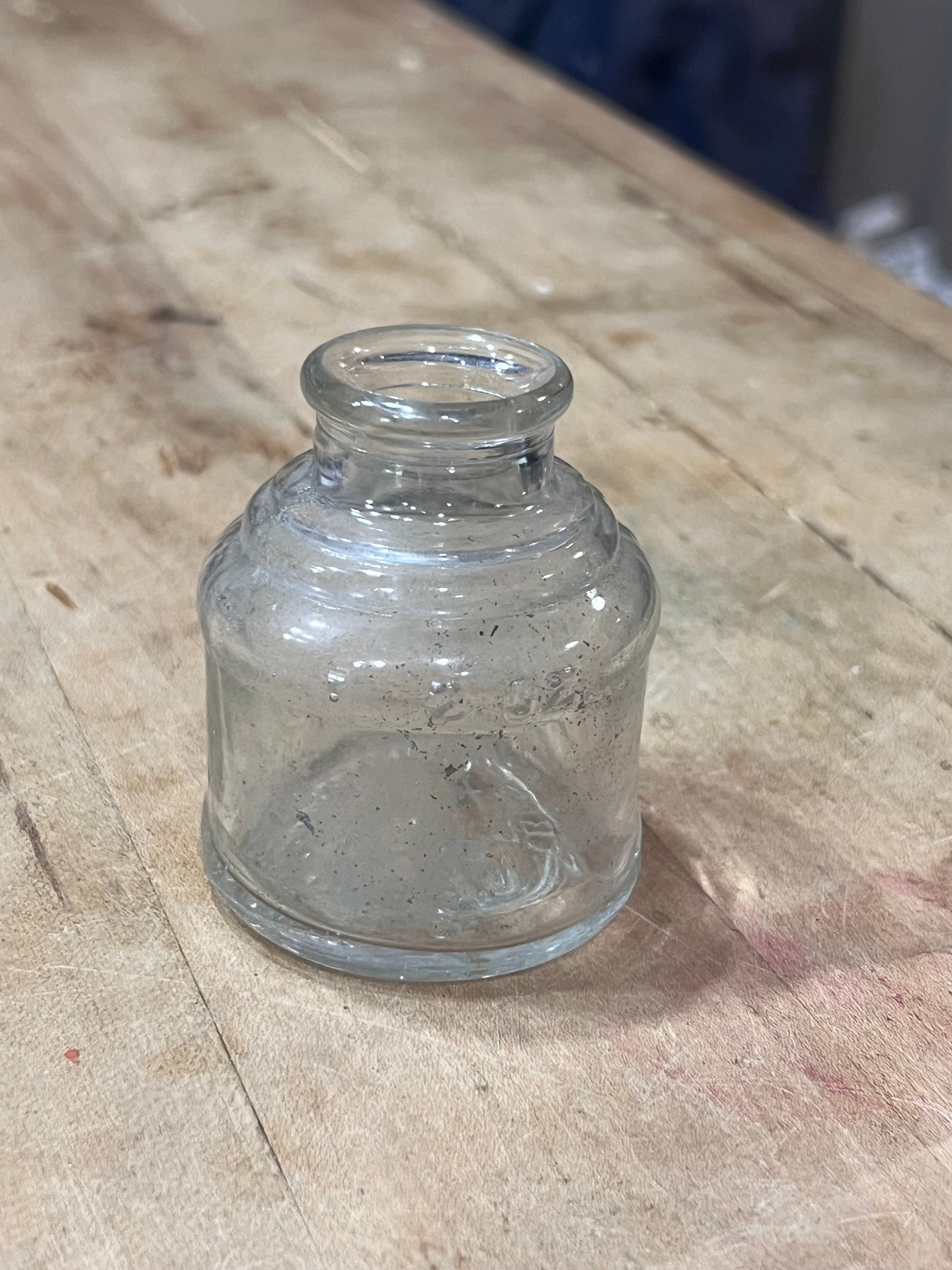A small, empty clear glass jar with a ribbed shoulder and a short neck, lying on a worn wooden surface.