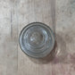 A clear glass bottle stopper with concentric circular ridges and a flared rim, shown from a top-down view on a light-colored wooden surface.